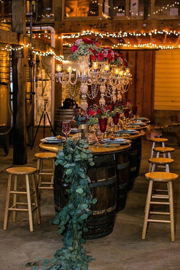 In questa foto un allestimento di nozze autunnale a tema vino realizzato in una cantina con  botti di legno usati come tavoli. Come centrotavola sono usati candelabri  colore argento con cristalli decorati con fiori rossi e lilla poggiati su un lungo runner di foglie di eucalipto. La mise en place riprende gli stessi colori con calici rossi. Vicino alle botti sono disposti sgabelli di legno chiaro