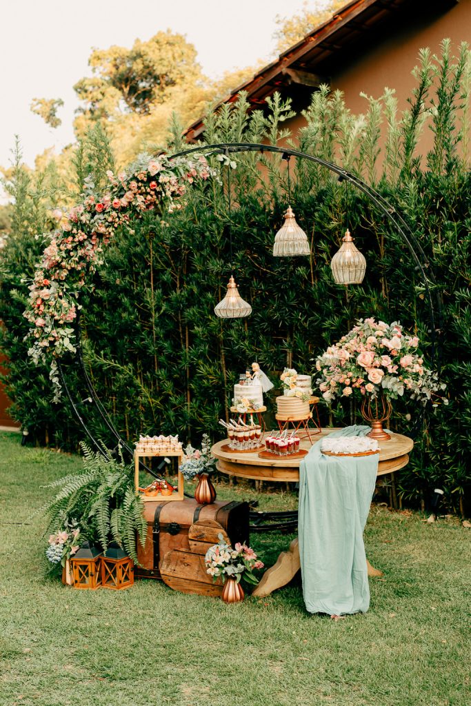 In questa foto uno sweet table allestito in un giardino . Su un tavolo di legno vintage e rotondo sono disposti dessert in bicchiere e torte. Una torta è posizionata su un drappo di stoffa verde salvia vicino a un vaso di colore bronzo con fiori rosa cipria e bianchi e foglie. Alla base è presente un vasetto di fiori, un baule vintage, lanterne di legno, una felce e un'alzata quadrata di legno con altri dolci al cucchiaio. Lo sweet table è sormontato da un arco di ferro decorato con fiori da cui pendono lampade vintage