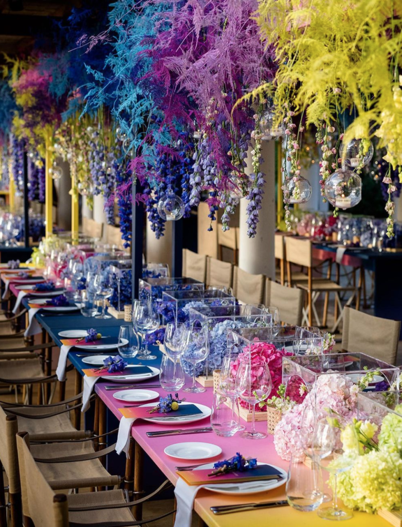 In questa foto l'allestimento di un ricevimento di nozze a gradazioni di colore blu, rosa, giallo e viola realizzato con sfere di vetro candele, piume e fiori pendenti nei colori abbinati e piccole composizioni disposte in fila al centro del tavolo