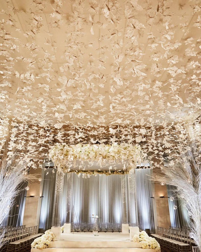 In questa foto allestimenti per un matrimonio a tema farfalle Total White. Il tetto è ricoperto di farfalle trasparenti. Ai lati sono disposti rammetti bianco ghiaccio a cui sono appese palline di trasparenti illuminate da candele. In basso è presente un altare per matrimonio civile quadrato circondato da fiori bianchi e con un baldacchino di farfalle. Ai lati sono presenti le sedie trasparenti per gli ospiti