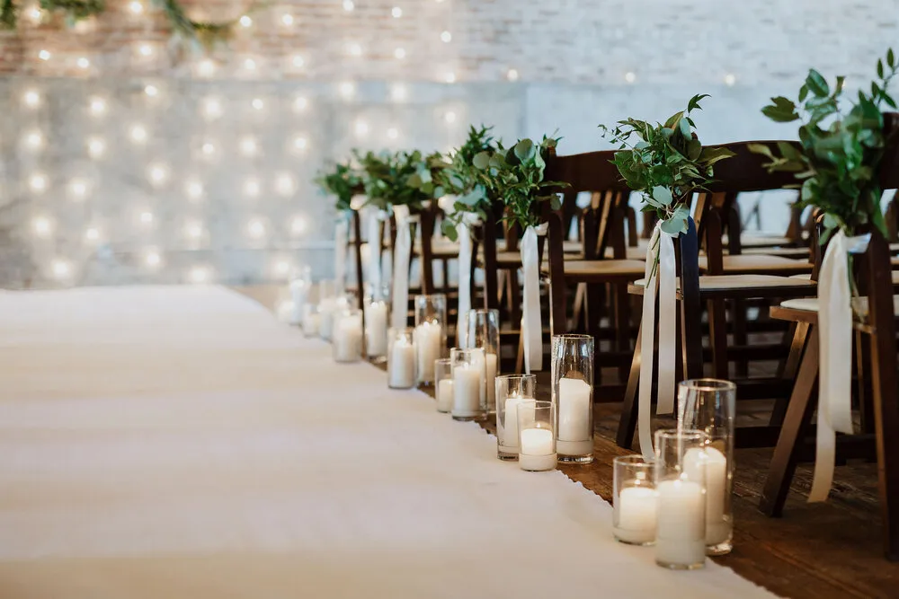 In questa foto l'allestimento della navata centrale della location di un matrimonio civile realizzato con un tappeto bianco lungo il quale sono disposti portacandele trasparenti con candele di diverse altezze. Sui banchi sono legati rametti d'alloro con nastri bianchi. Sul fondo si intravede una parete di luci