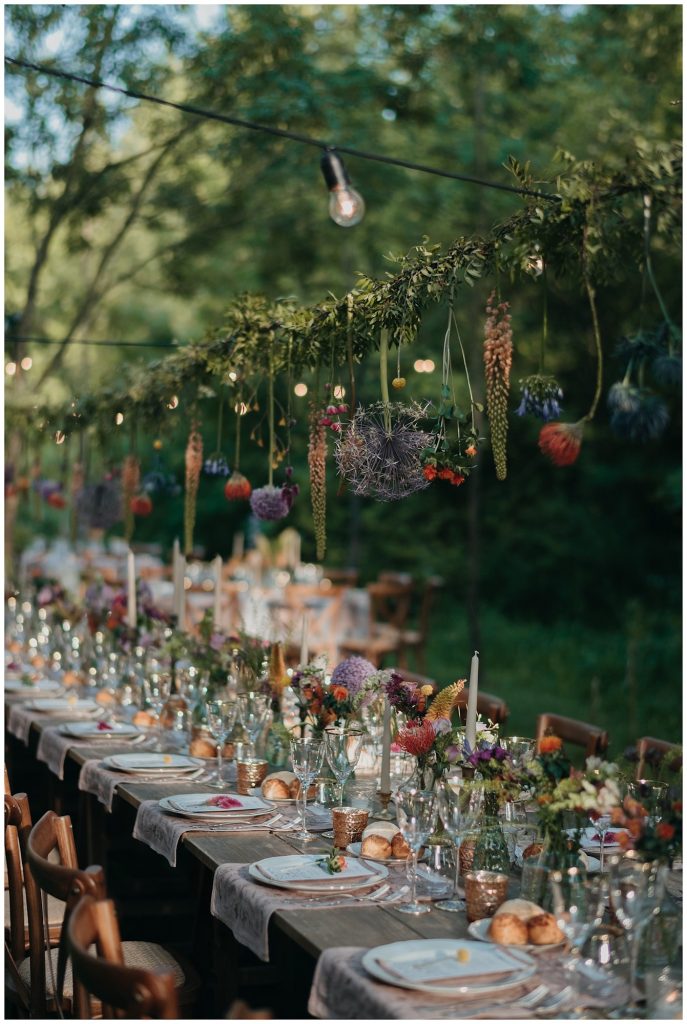 In questa foto l'allestimento di un ricevimento di nozze in stile Rustic Chic realizzato in un bosco con tavoli imperiali di legno grezzo decorati con vasetti di fiori di campo intrecciati anche su una struttura pendente 