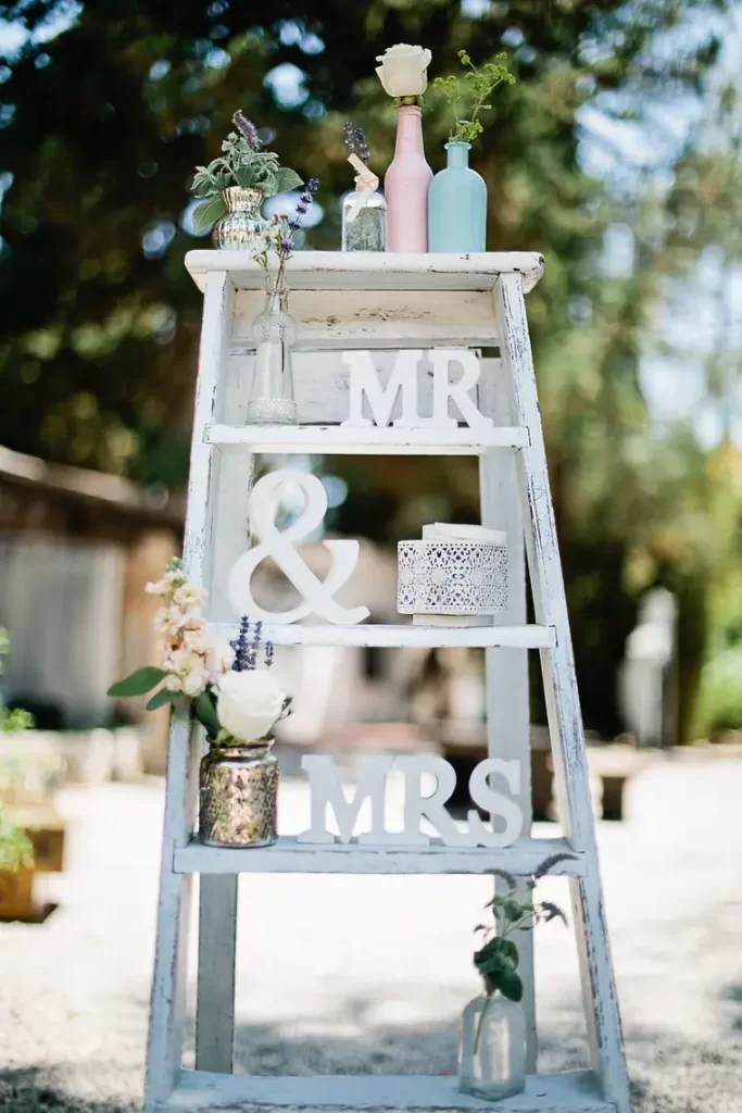 In questa foto una scala bianca in stile Shabby Chic inserita tra gli allestimenti di un matrimonio in un giardino. Sulla scala sono disposti in ordine dalla cima: 4 vasetti di argento, trasparente, rosa e azzurro con fiori di campo; sui gradini una scritta con "Mr & Mrs" e vasetti con altri fiori di campo