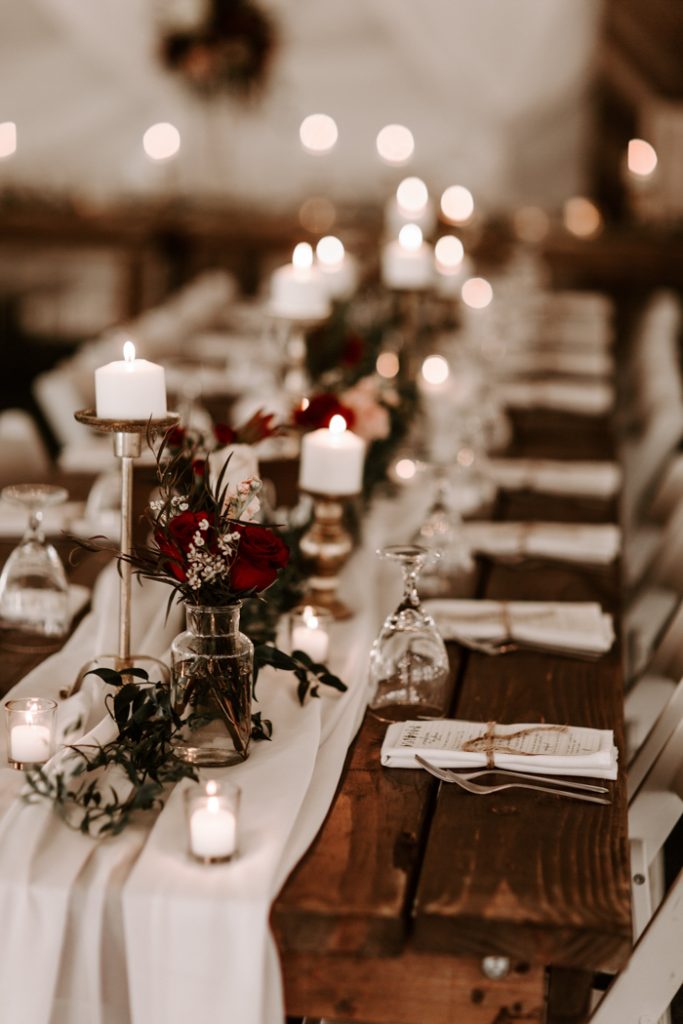 In questa foto l'allestimento di un tavolo per matrimonio invernale nei toni del bianco e del rosso. Il tavolo di legno nudo è decorato con un runner di stoffa bianca su cui sono adagiate piccole candele bianche, candelabri colore argento con una candela bassa bianca, vasetti di vetro con rose rosse e nebbiolina. Ad ogni tovagliolo è legato un menù. Completa la decorazione rametti di agrifoglio intrecciati al runner