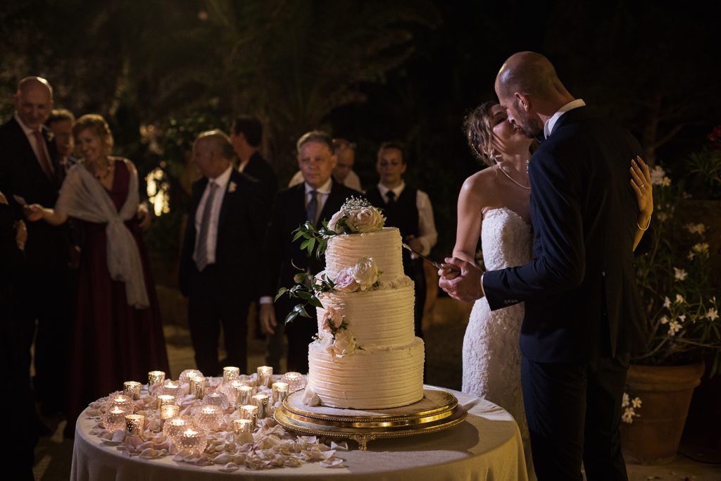 In questa foto due sposi si baciano mentre tagliano la torta nuziale bianca decorata con fiori rosa. Il tavolo della torta è decorata con piccoli portacandele a forma di boll. Dietro si intravedono gli ospiti