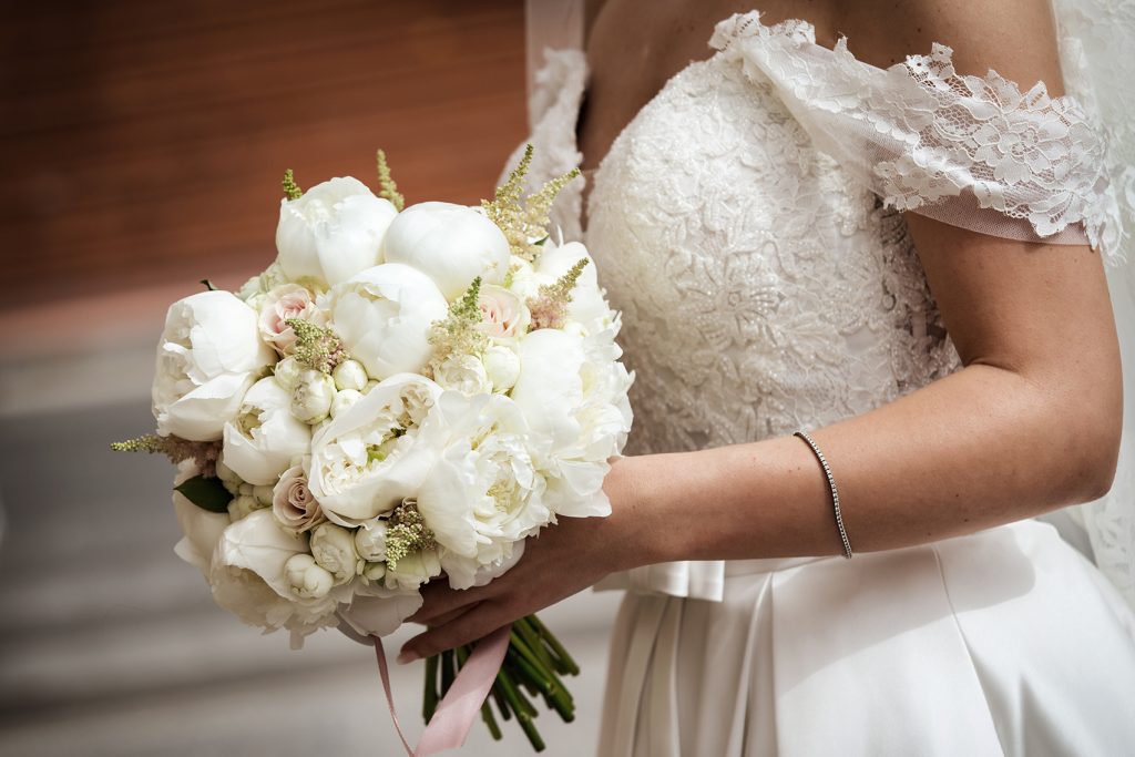 In questa foto il dettaglio di un bouquet di peonie bianche e piccole rose rosa tenuto in mano dalla sposa