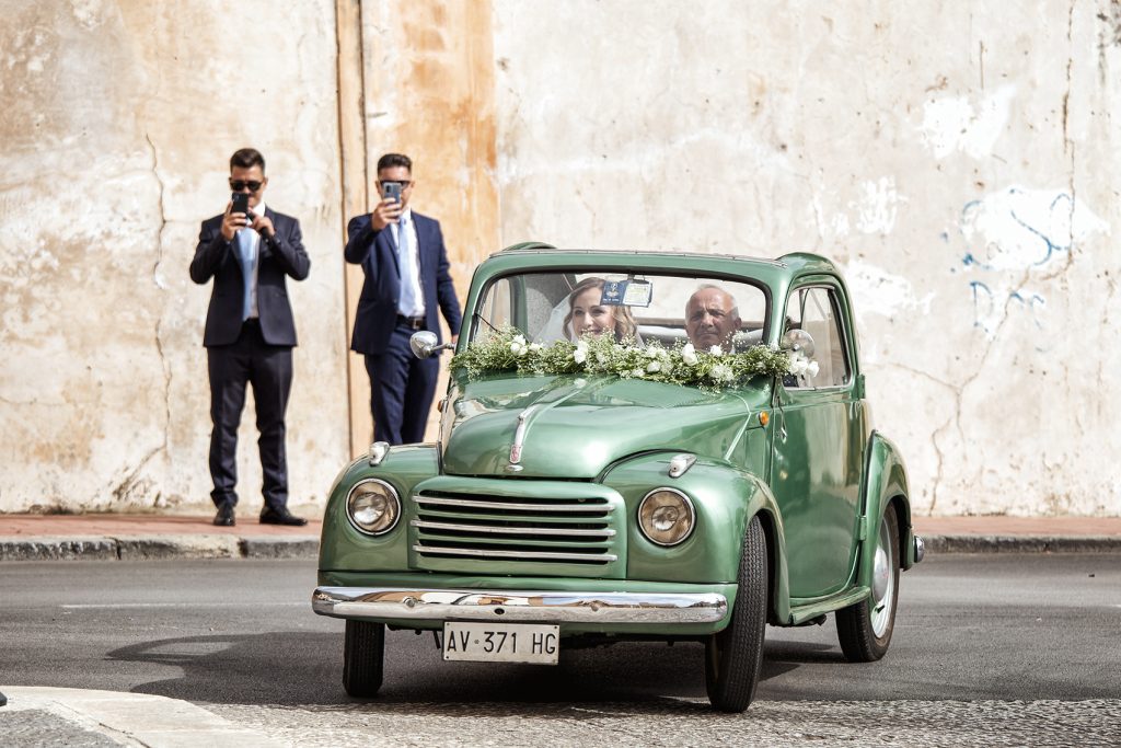 In questa foto una sposa arriva con il papà a bordo di un'auto d'epoca verde chiaro decorata  sul parabrezza con nebbiolina e foglie