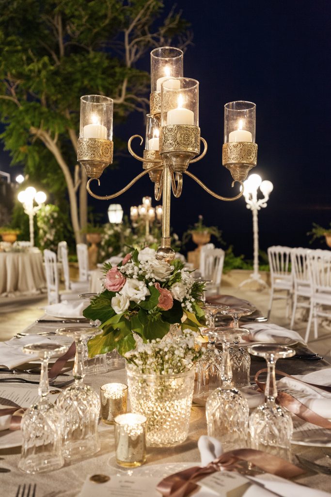 In questa foto il dettaglio della mise en place su un tavolo imperiale nei toni del bianco e del rosa. Al centro è presente un piccolo mazzetto di rose rosa e bianche in un vaso di cristallo. Dietro si intravede un candelabro dorato con candele accese