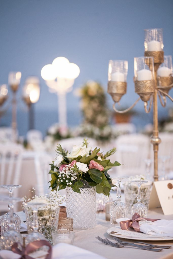 In questa foto il dettaglio della mise en place su un tavolo imperiale nei toni del bianco e del rosa. Al centro è presente un piccolo mazzetto di rose rosa e bianche in un vaso di cristallo. Dietro si intravede un candelabro dorato con candele 