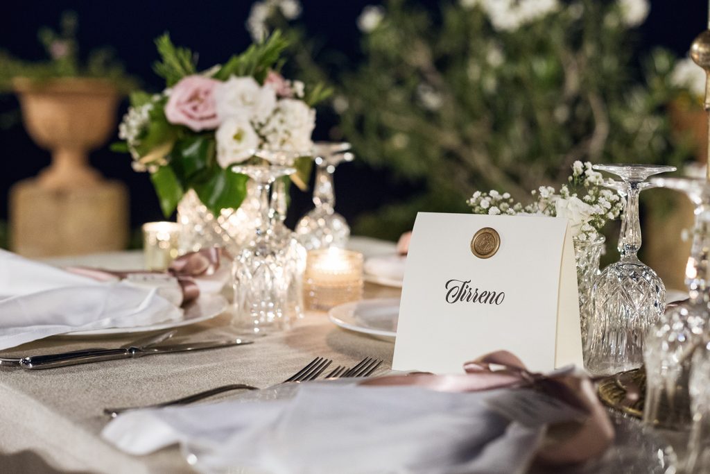 In questa foto il dettaglio di una mise en place nei toni del bianco e del rosa. Sul tavolo è presente una targhetta con il nome del tavolo a tema mare. Sulla targhetta è scritto "Tirreno" ed è presente u bollino di ceralacca colore oro