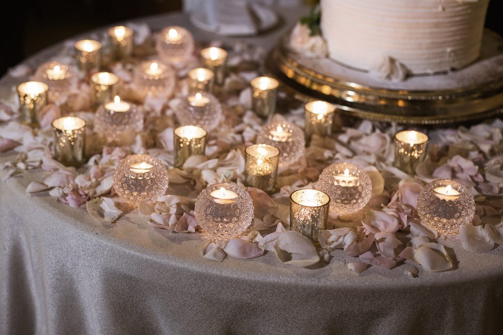 In questa foto un dettaglio della decorazione del tavolo della torta nuziale con petali rosa e piccoli portacandele di vetro e di forma sferica con candele accese.