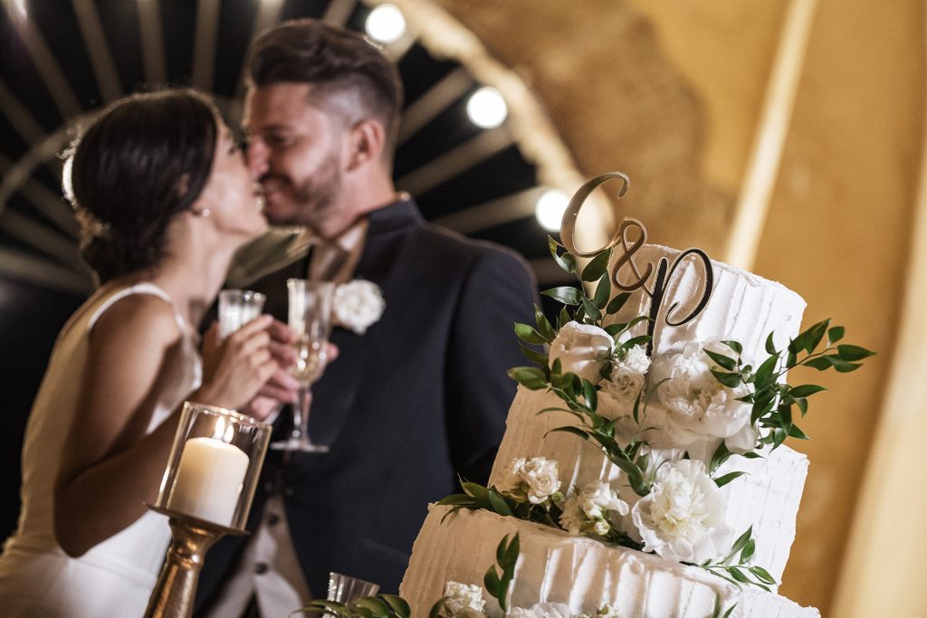 In questa foto due sposi in secondo piano si baciano sorridenti facendo un brindisi dietro la torta nuziale con le iniziali dei loro nomi G e P. La torta ripresa in obliquo è decorata con fiori bianchi e foglie