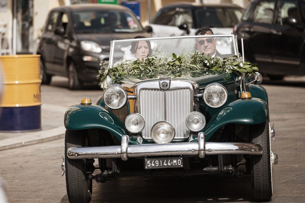 In questa foto una sposa arriva con il papà a bordo di un'auto d'epoca verde scuro decorata con nebbiolina e foglie