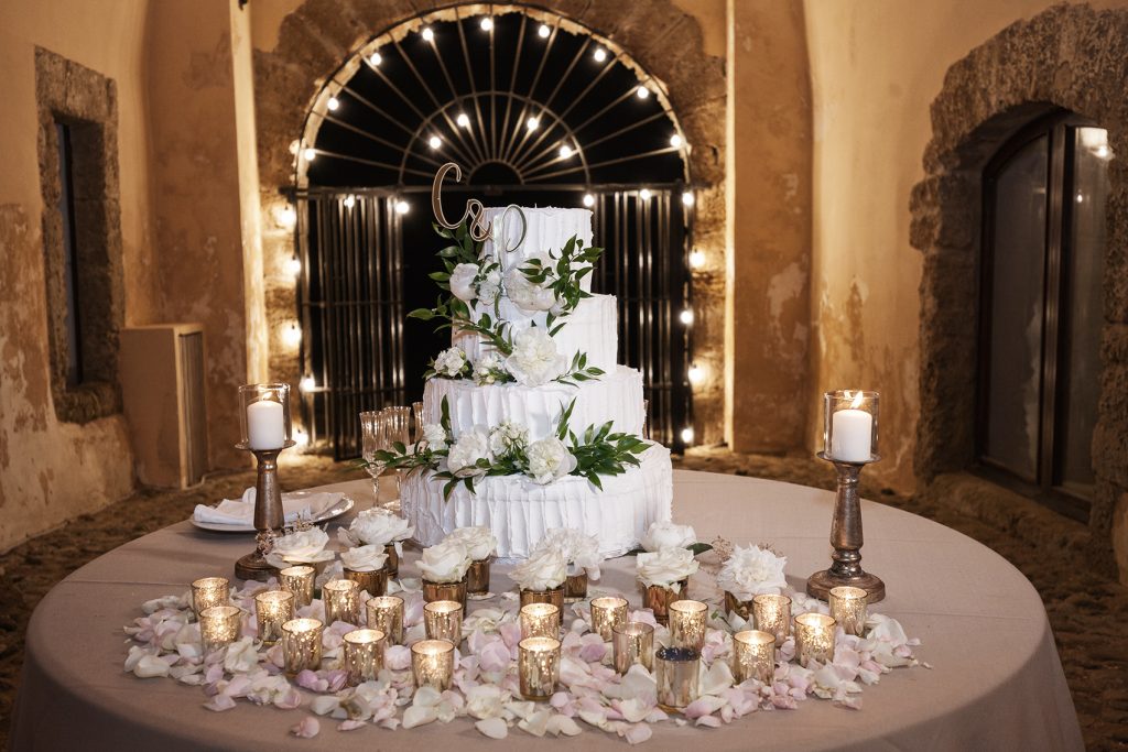 In questa foto una torta nuziale bianca decorata con fiori bianchi e foglie. Davanti al tavolo sono presenti petali di rose e piccoli portacandele colore oro