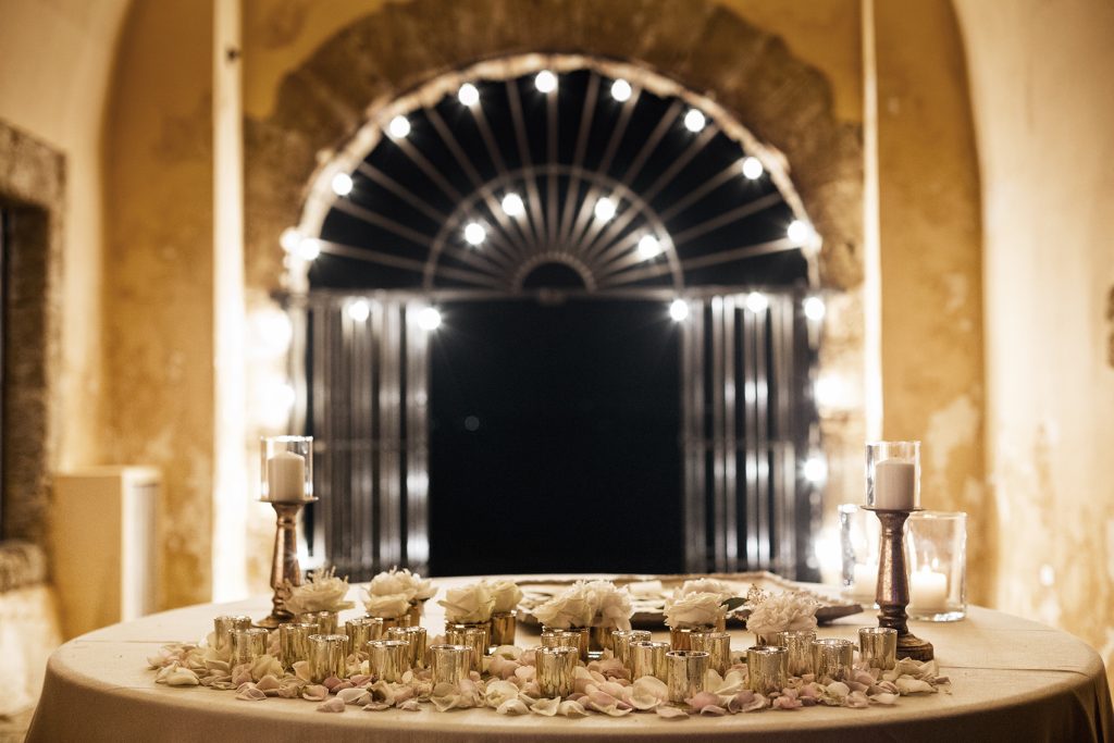 In questa foto un tavolo con portacandele colore argento con petali di rose. Sullo sfondo si vede un arco illuminato da piccole luci