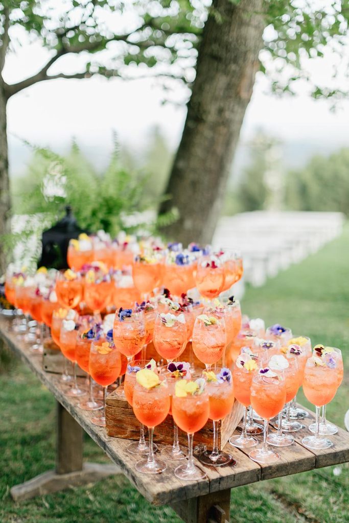 In questa foto il tavolo di legno con alzatine abbinate per un ricevimento di nozze su un prato allestito dal catering  sotto un albero per l'aperitivo con cocktail arancioni in calici trasparenti decorati con fiori colorati di campo