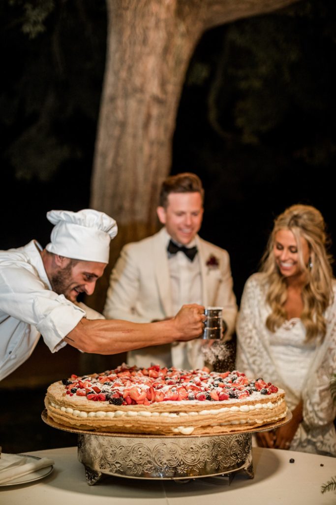 In questa foto il pasticcere del catering del matrimonio prepara e decora dal vivo la torta nuziale millefoglie con panna e frutti di bosco mentre gli sposi, in secondo piano e fuori fuoco, guardando sorridenti