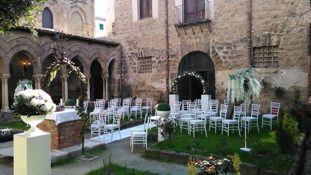 In questa foto il chiostro della Magione a Palermo decorato per un matrimonio all'aperto con alzatine di fiori bianchi, archi di fiori, sedie bianche e un tappeto bianco