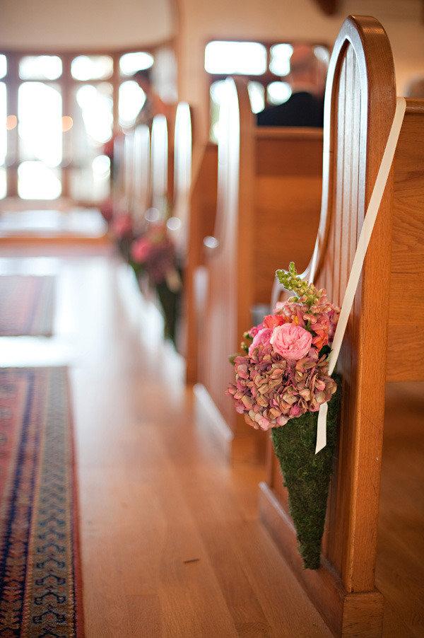 In questa foto in primo piano un cono di muschio verde ripieno di fiori rosa e verdi, legato al banco di una chiesa con un nastro sottile di seta. Fuori fuori, dietro, si intravedono altri coni dello stesso tipo. Lungo il bordo sinistro della foto ci sono dei tappeti