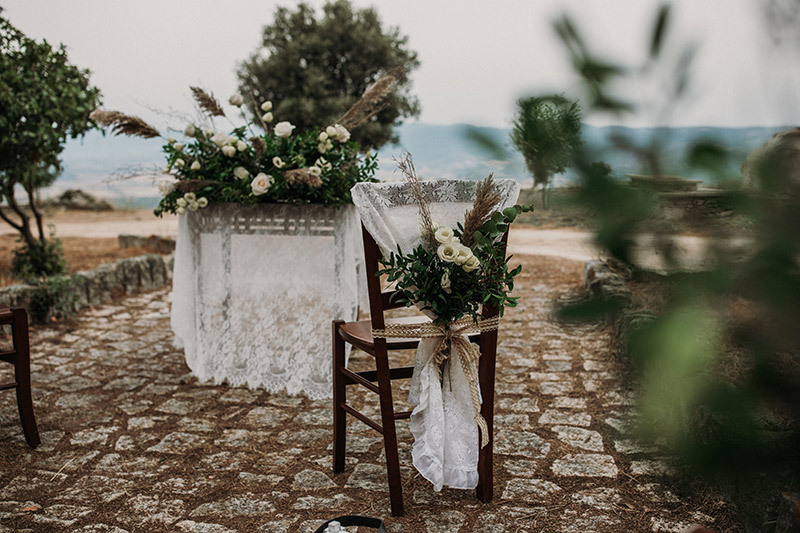 In questa foto gli allestimenti del rito civile all'aperto. In primo piano una sedia di legno, molto rustica, ricoperta da un tessuto ricamato. A questo è legato un mazzo di fiori, con rami di eucalipto e altre piante selvatiche e ranuncoli bianchi. A tenerli insieme un nastro di juta. in secondo piano un tavolo ricopero dallo stesso tessuto ricamato e sopra una rigogliosa composizione floreale, tra piante selvatiche verdi, rose e ranuncoli bianchi e pampas. 