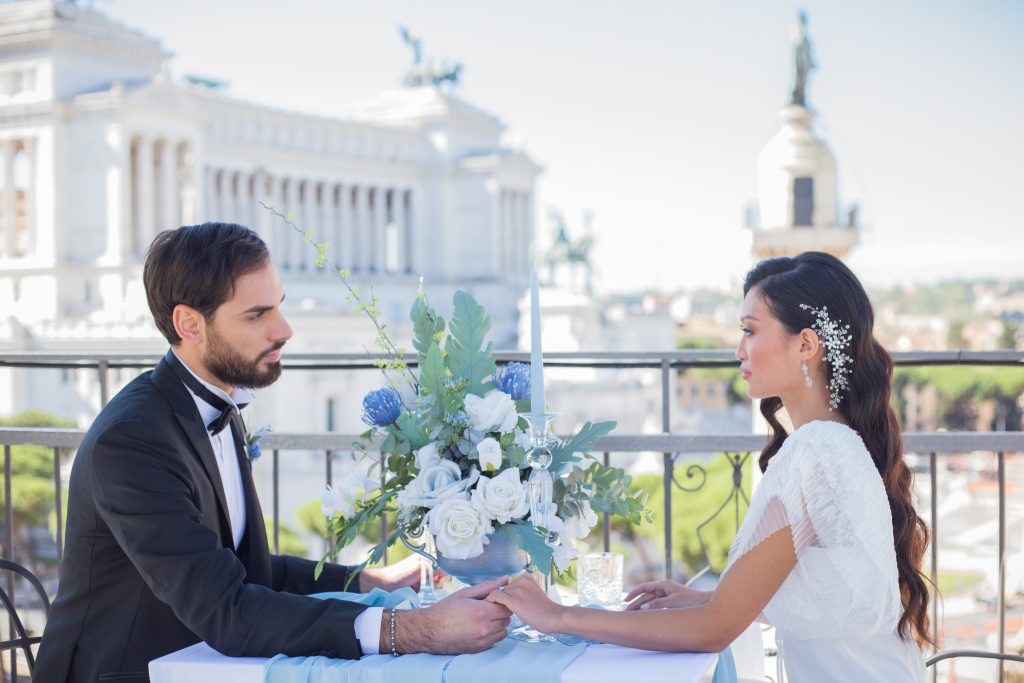 In questa foto una coppia di sposi si tiene per mano seduta ad un tavolo allestito per le nozze nella terrazza dell'NH Collection Roma Fori Imperiali, tappa del Wedding Tour NH Collection