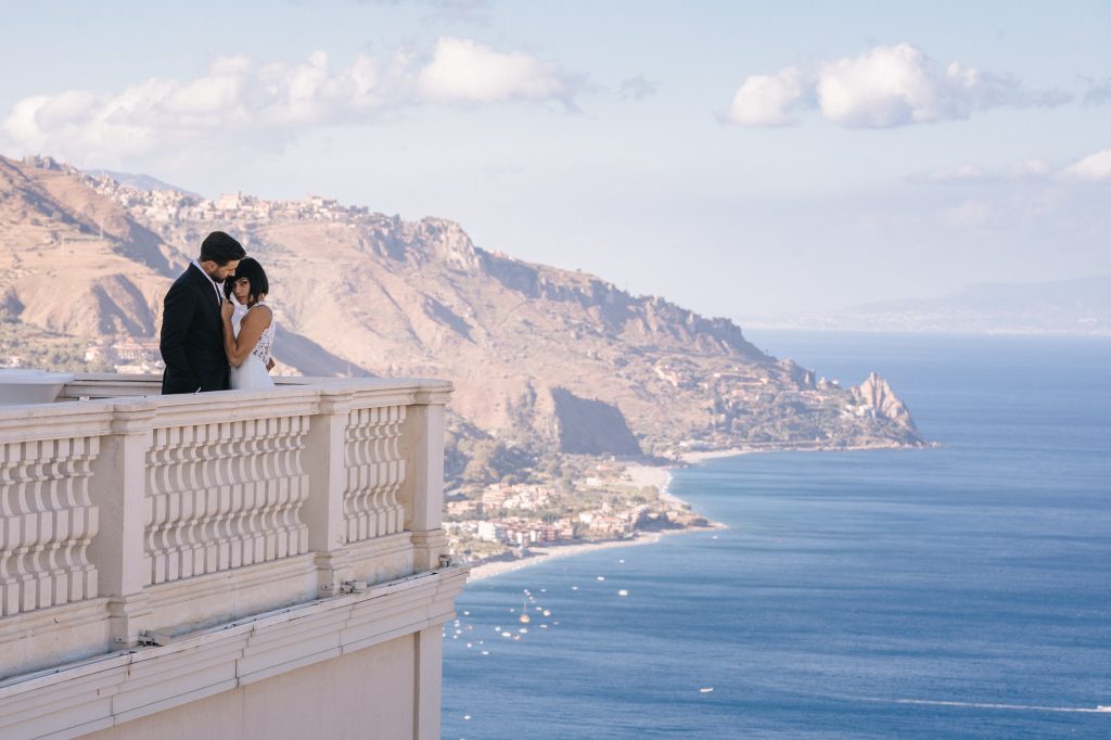 In questa foto due sposi si abbracciano sulla terrazza dell'NH Collection Taormina, che si affaccia sul panoramico e mozzafiato golfo siciliano; l'hotel sarà una delle tappe del Wedding Tour NH Collection 