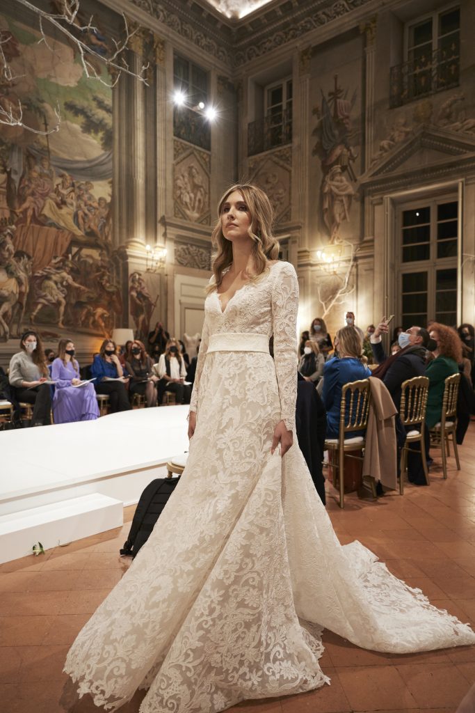 In questa foto una modella sfila indossando un abito da sposa interamente in pizzo, con gonna scivolata e scollo a V, della capsule collection Revenge Atelier 2022