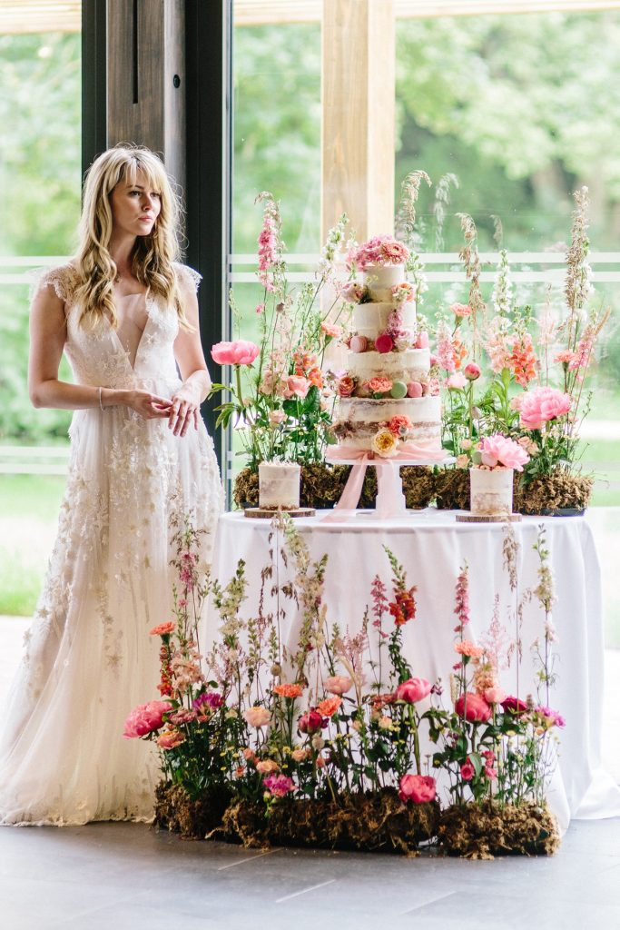 In questa foto una sposa accanto al tavolo allestito per la torta nuziale con fiori di campo alla base, sul tavolo e sui piani della torta 