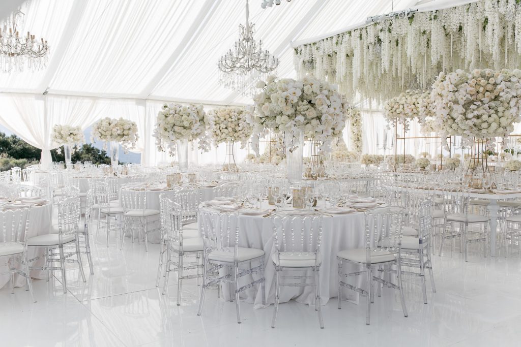 In questa foto l'allestimento di un ricevimento di nozze Total White. All'aperto, sotto una tenda, sono disposti tavoli rotondi con sedie chiavarine trasparenti. Al centro di ogni tavolo è presente un'alazata trasparenti con bouquet di fiori bianch. Sul tavolo degli sposi a destra è presente un tetto di fiori pendenti bianchi. Completano l'allestimento lampadario di cristallo