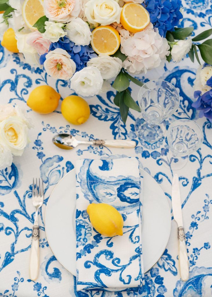 In questa foto il dettaglio di una mise en place blu, bianca e gialla ispirata alla Costa Amalfitana. La tovaglia e il tovagliolo sono decorati con il tema delle maioliche. Un limone è usato come segnaposto e nel centrotavola di fiori bianchi, rosa e blu. La mise en place è completata con posate d'argento