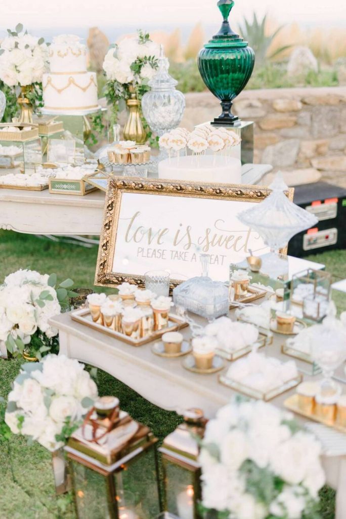 In questa foto uno sweet table per un matrimonio a tema bianco e oro.Lo sweet table è composto da 2 tavoli uno davanti all'altro. In primo piano è inquadrato un tavolo con dolci al cucchiaio in coppette colore oro disposti su vassoi e coppe di cristallo. Il tavolo è decorato con uno specchio con cornice dorata con su scritto Love is Sweet. Dietro è presente un'altro tavolo con altri dolci e torte