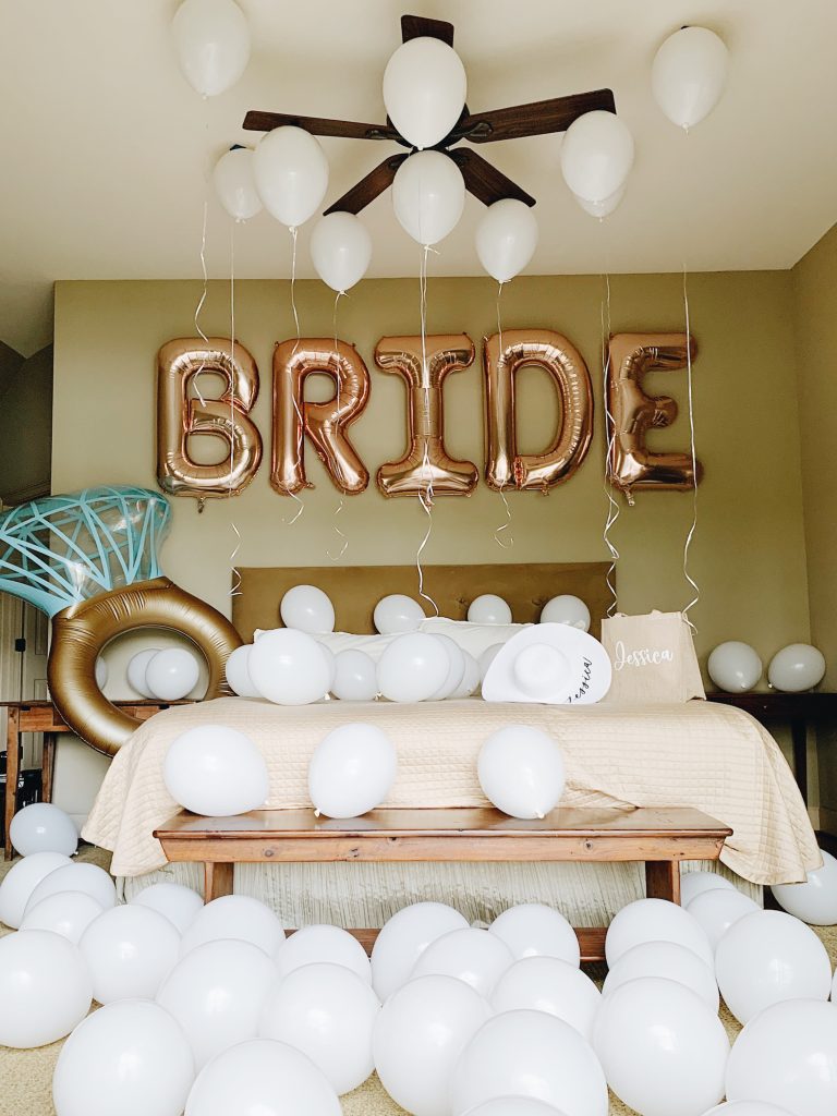 In questa foto un camera da letto addobbata con un palloncino colore oro con scritto Bride, palloncini bianchi sul pavimento e sul letto e un altro palloncino a forma di anello posto sull'angolo sinistro 