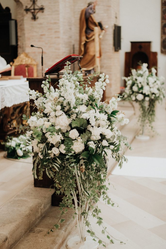 In questa foto allestimenti matrimonio floreali posti ai lati dell'altare di una chiesa realizzati con garofani, rose e gladioli bianchi e rami di alloro