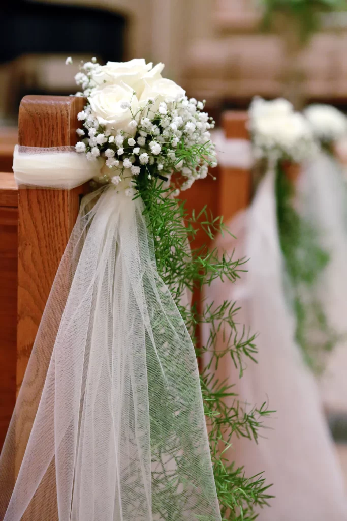 In questa foto un dettaglio delle decorazioni per i banchi di una chiesa per matrimonio realizzate con piccoli bouquet di rose bianche, nebbiolina, felce e nastri di tulle bianchi