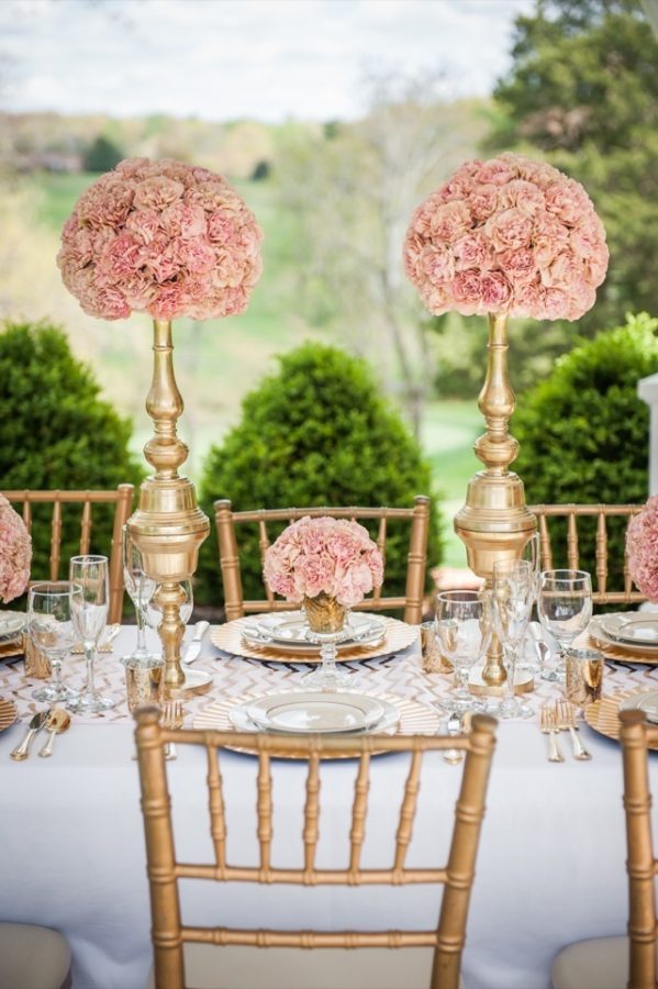 In questa foto una mise en place per un matrimonio elegante nei toni dell'oro e del rosa. Il tavolo è fotografato lateralmente con le sedie chiavarine colore oro in primo piano. Sul tavolo sono presenti candelabri con composizioni di garofani rosa. Le stesse composizioni sono poggiate sui piatti come segnaposti