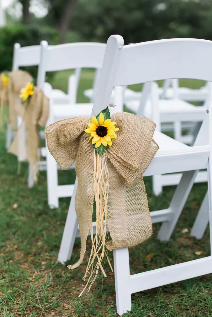 In questa foto il dettaglio dell'allestimento di un matrimonio civile su un prato, realizzato con un fiocco di iuta decorato con un girasole e paglia fissato ad una sedia per la cerimonia