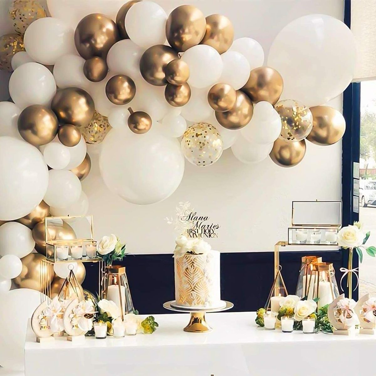 In questa foto una scultura di palloncini bianchi e colore oro fa da sfondo ad un tavolo su cui è presente, al centro ,una torta nuziale su un'alzata e con una scritta dorata sulla sommità. Mentre sui lati sono presenti candele basse e altre decorazioni bianche e dorate 