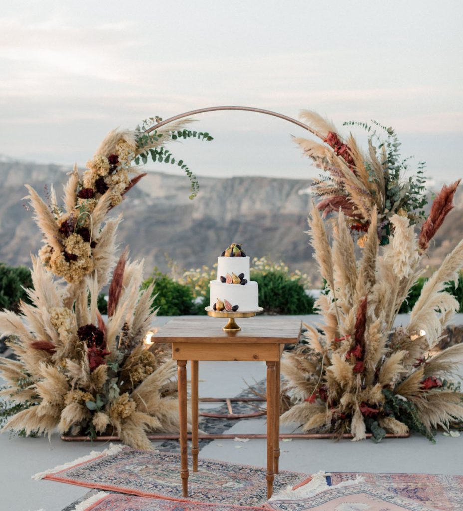 In questa foto un piccolo tavolo di legno su cui è poggiata la torta nuziale decorata con piccole pere. Dietro il tavolo è presente un arco addobbato con pampas beige, rosse e verdi. Completano l'allestimento alcuni tappeti 