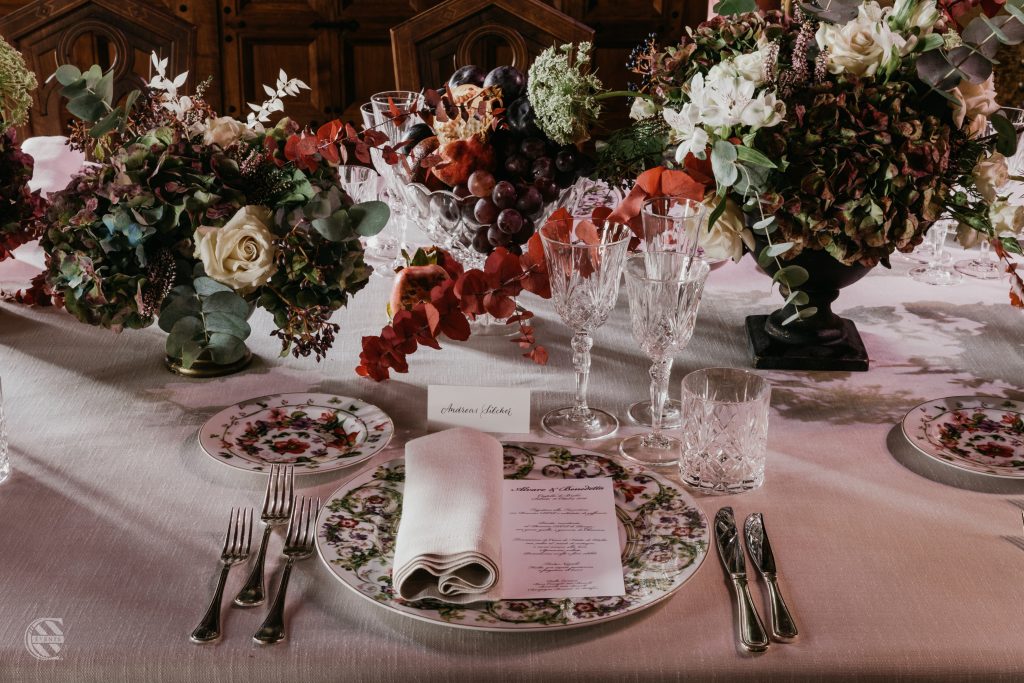 In questa foto una raffinata mise en place a tema autunnale con piatti decorati, bicchieri in cristallo e centro tavola con fiori e frutta di stagione.