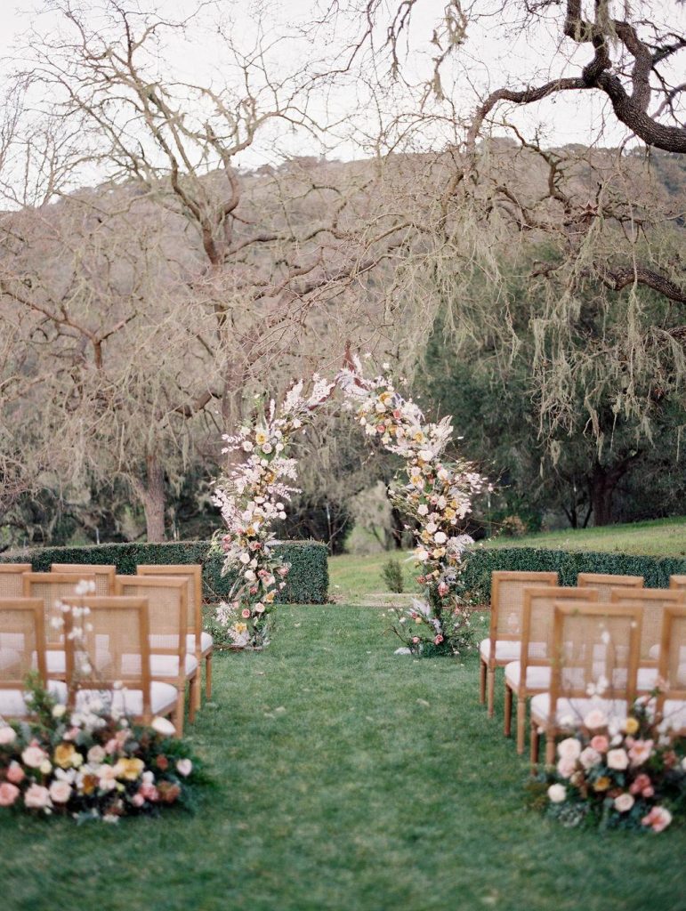 In questa foto l'allestimento di un matrimonio civile in giardino sotto un albero senza foglie. Dietro l'ultima fila di sedie di legno chiaro sono disposte due composizioni di fiori rosa, gialli e bianchi. In fondo al corridoio centrale è presente un arco con gli stessi fiori