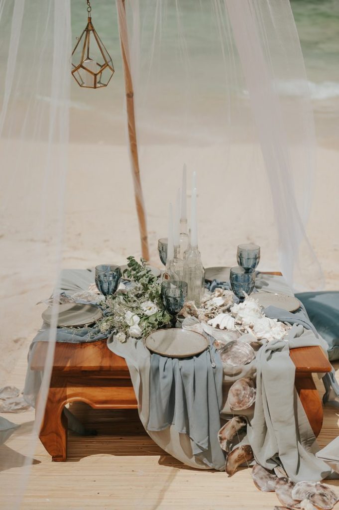 In questa foto il dettaglio di una mise en place nei toni dell'azzurro e del grigio allestita su un tavolo basso su una spiaggia circondato una tenda di tulle. Sul tavolo sono presenti drappi di stoffa blu azzurra e grigia su cui sono poggiati piatti grigi, calici blu, fiori bianchi, conchiglie intrecciate e un centrotavola di candele bianche inserite in bottiglie di vetro