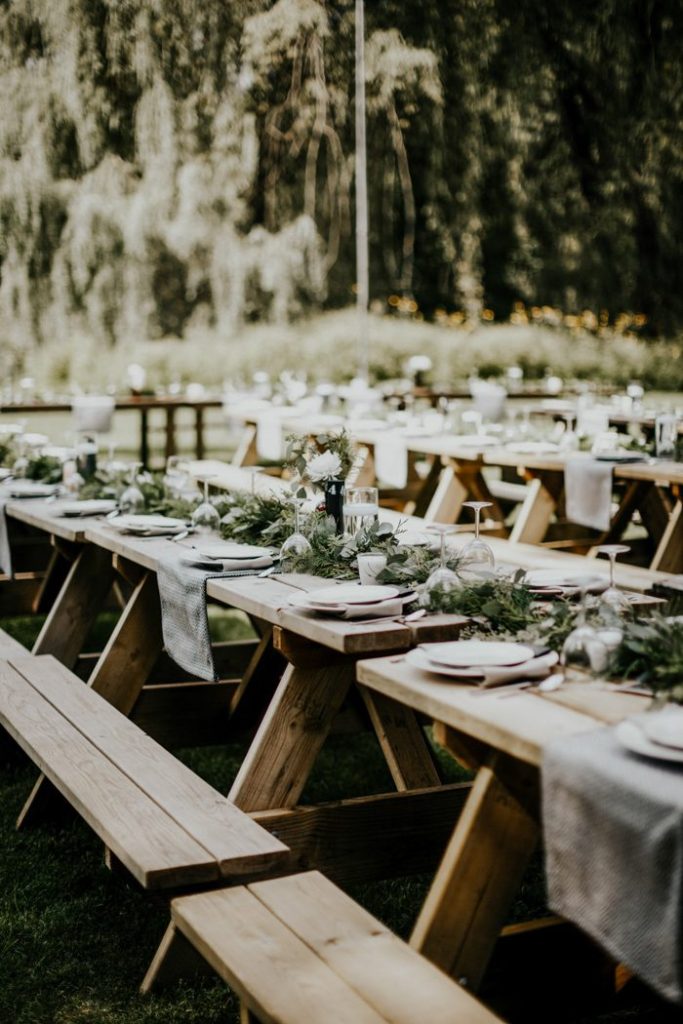 In questa foto un ricevimento di nozze allestito in un bosco su tavoli con panchine in legno inquadrati in obliquo da destra verso sinistra, decorati con tovaglioli azzurro polvere e un runner di foglie e fiori bianchi 