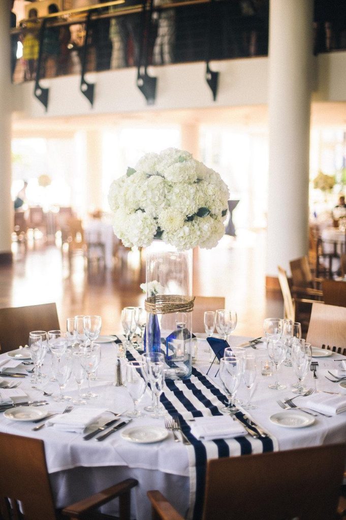 In questa foto un tavolo matrimonio rotondo con un runner a righe bianche e blu. A centrotavola un vaso lungo trasparente con fiori bianchi