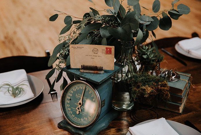 In questa foto un centrotavola per un matrimonio a tema vintage realizzato con un'antica bilancia, foglie di eucalipto, una cartolina postale e un pettine vintage