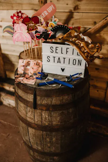 In questa foto un angolo photoboothe con accessori per selfie poggiati su una botte di legno. È presente anche una tag con scritto "Selfie Station"