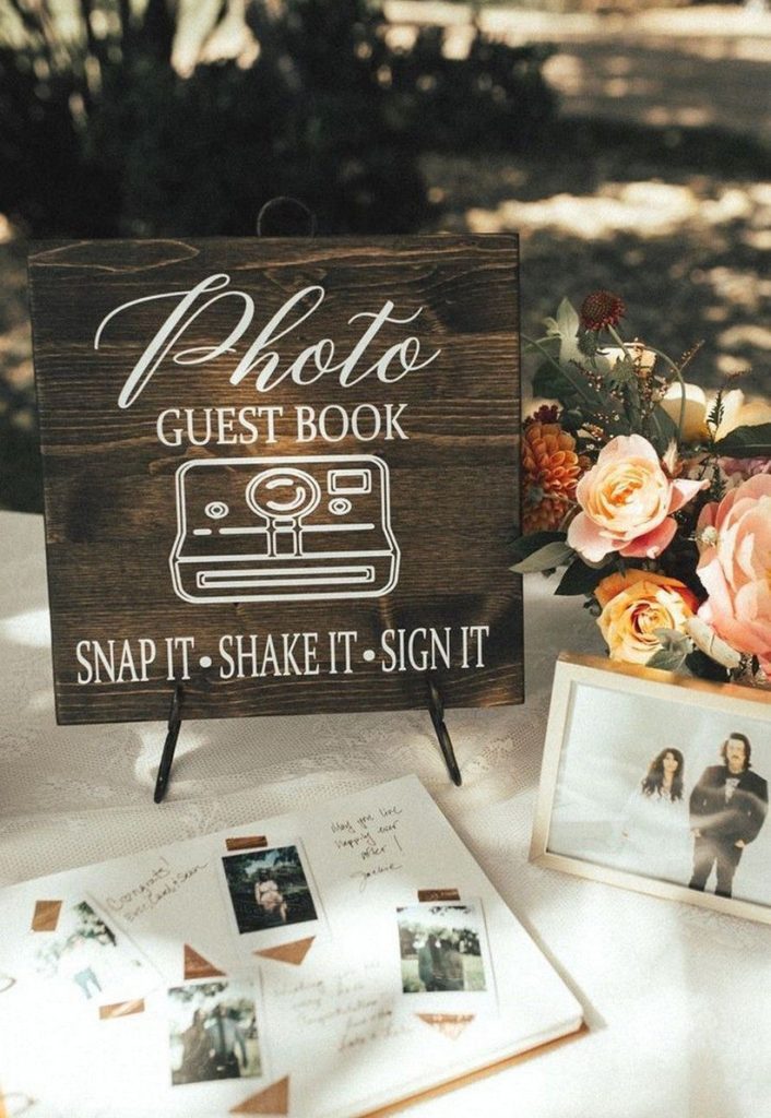In questa foto un tavolo guestbook allestito con una foto degli sposi, un album con le foto e i messaggi degli ospiti, dietro una targa di legno con scritto "Photo GuestBook" e un composizione di rose arancioni