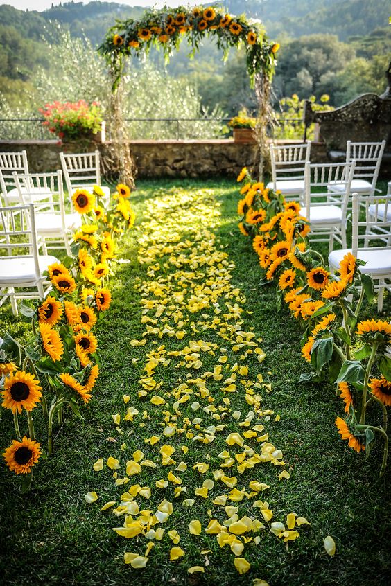 In questa foto la location di un matrimonio civile su un prato con i girasoli disposti lungo il corridoio centrale coperto di petali gialli. Accanto al corridoio centrale sono presenti sedie chiavarine bianche. In fondo, davanti ad un muretto di pietra, è presente un arco decorato con paglia e girasoli sul lato orizzontale