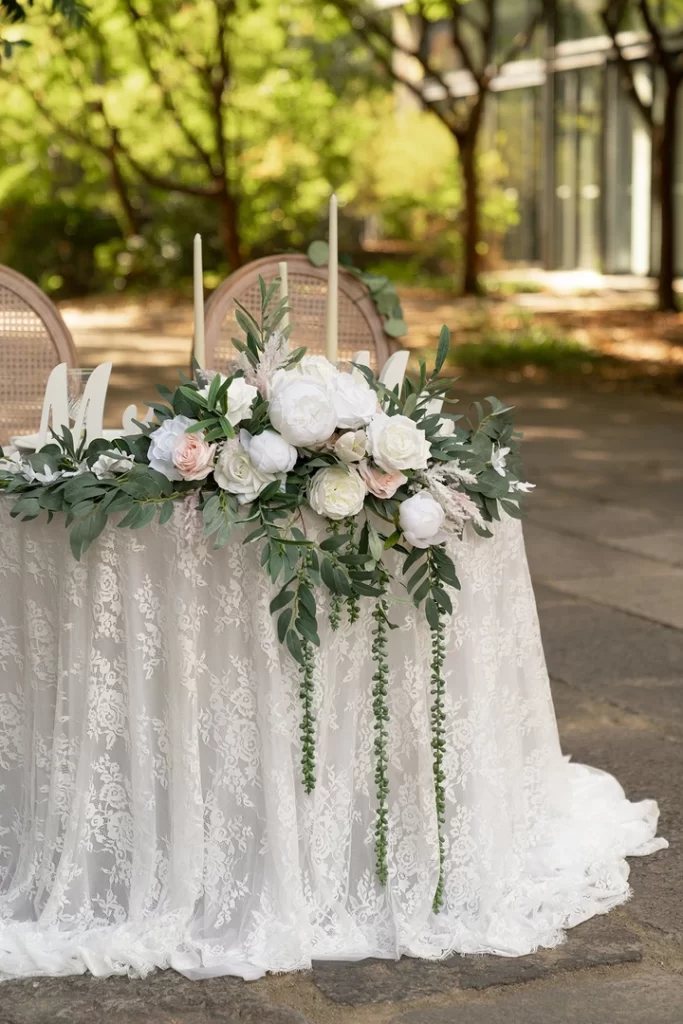 In questa foto una composizione di peonie bianche e rami di foglie pendenti circonda il bordo esterno del tavolo degli sposi decorato con una tovaglia di pizzo chantilly
