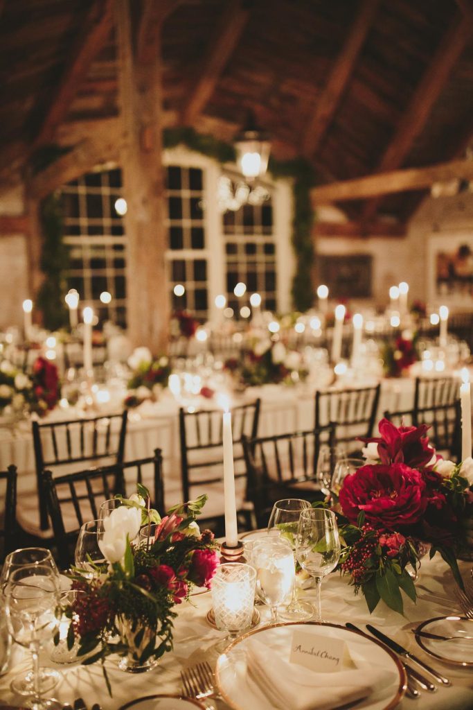 In questa foto una sala allestita per un   matrimonio natalizio con fiori rossi e bianchi. I tavoli usati sono imperiali disposti l'uno accanto all'altro. Completano la mise en place candele bianche lunghe accese, piatti di porcellana con bordo colore oro, posate in argento e calici di cristallo