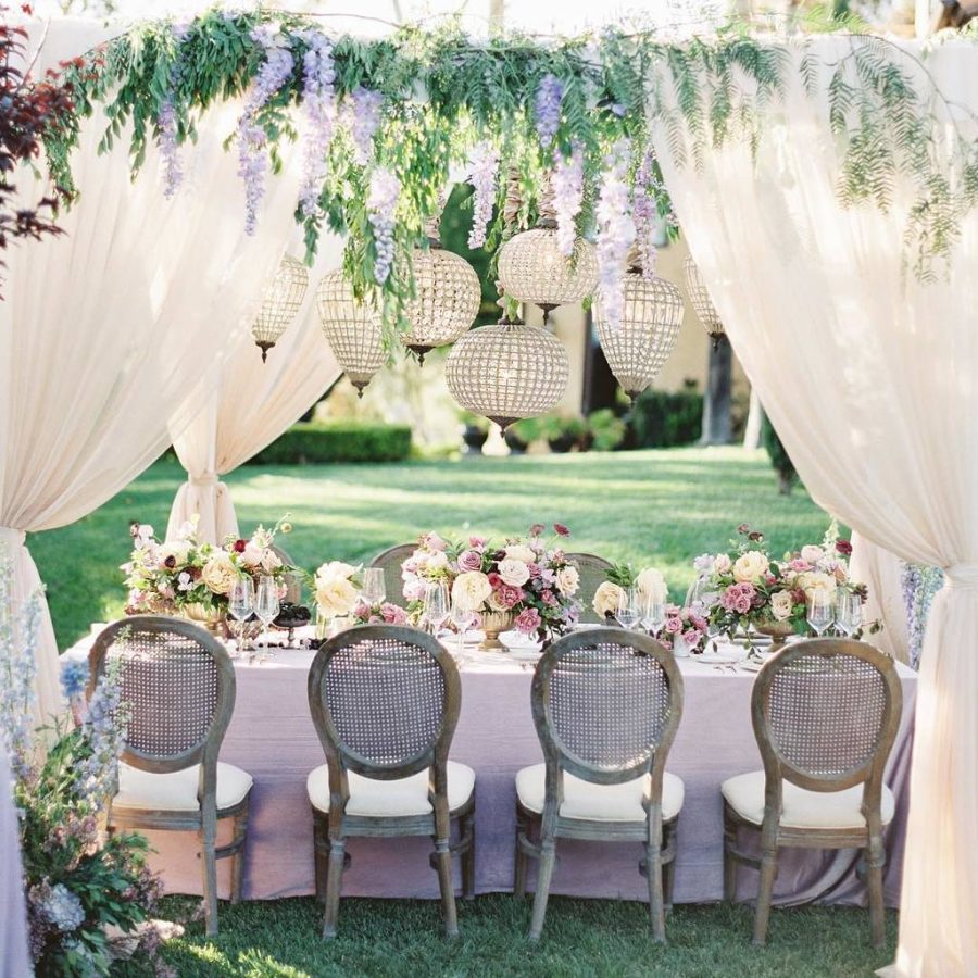 In questa foto un tavolo matrimonio allestito su un prato sotto tende bianche decorate con fiori lilla e foglie rampicanti. Sul tavolo è distesa una tovaglia colore lilla ed è decorato con centrotavola colore oro con rose bianche e lilla. Sopra il tavolo sono presenti lampadari rotondi di cristallo. Lungo il tavolo sono disposte quattro sedie a medaglione di legno inquadrate da dietro